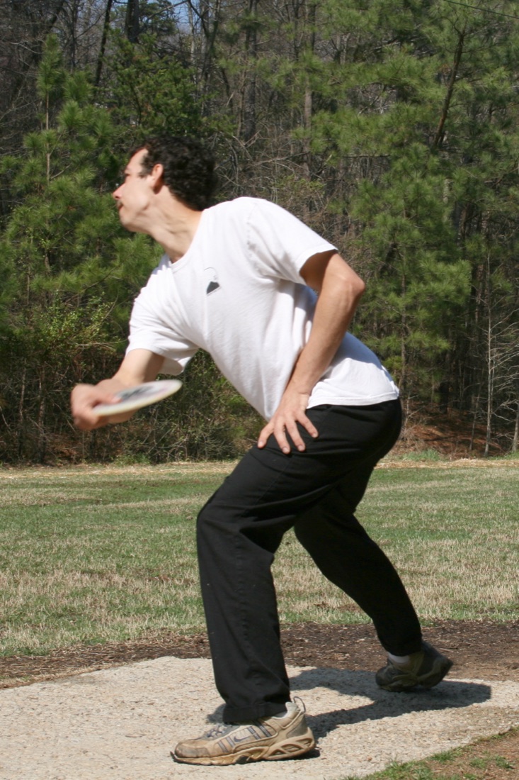 A disc golfer mid-throw, reaching back to release the disc on a wooded course